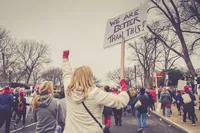 Group of protestors march down a street. One woman raises a sign reading, 'We are better than this'