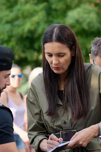 A woman in a crowd shows what she is writing on a piece of paper to a man watches with interest