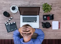 Top-down view of a man sitting at a table with a laptop in front of him. His hands are on his head, which is bowed and he is getting nothing done.