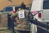 A note reading 'Register to vote here' is pinned to the back of a volunteer in a voter registration drive